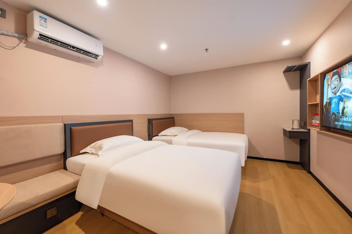 Ein Hotelzimmer mit zwei Betten, einer Bank, einer Klimaanlage, einem Fernseher und einem Regal mit einer Fernbedienung.