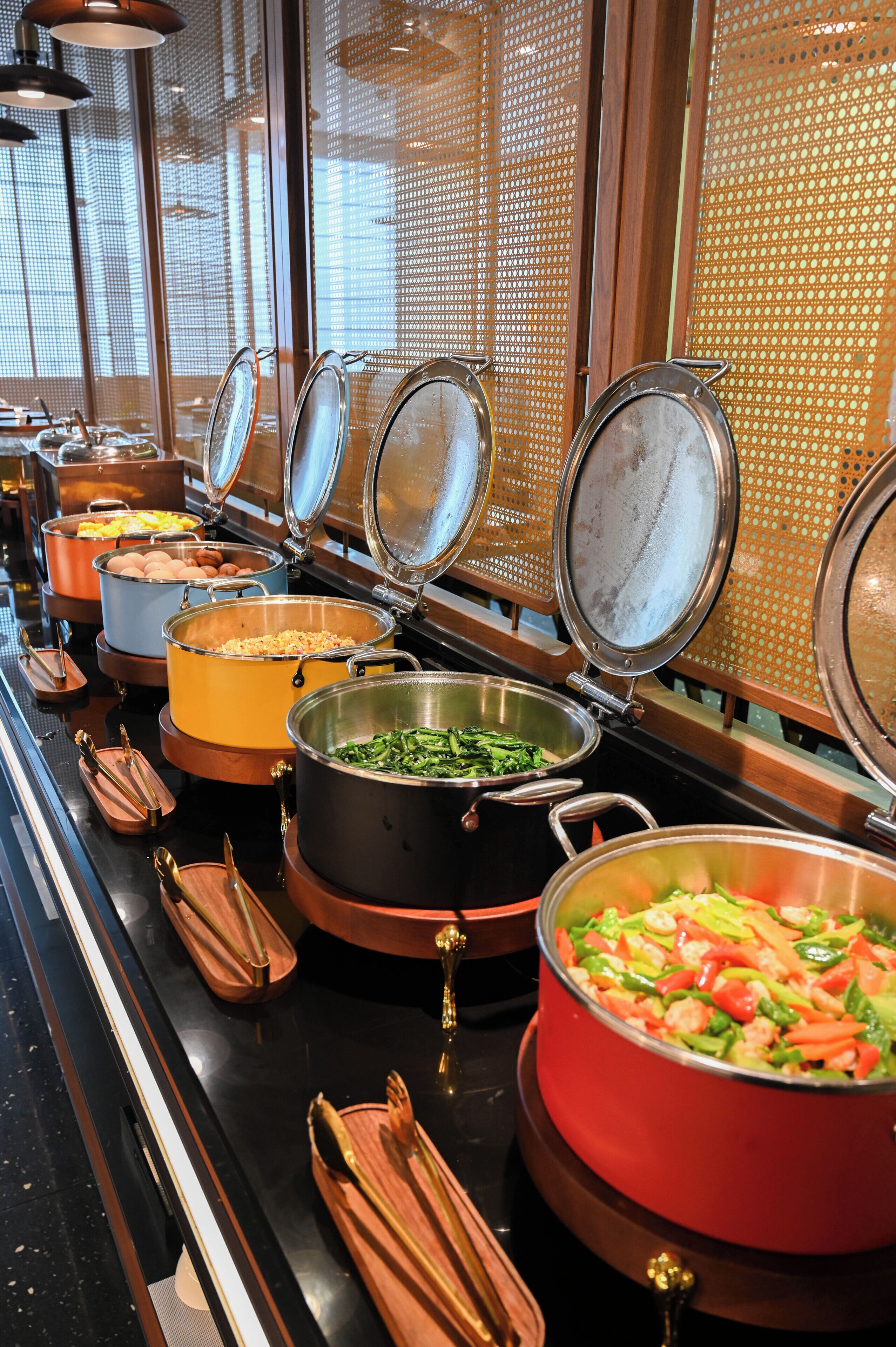 Tarjolla on runsas buffet, jossa on erilaisia ruokia suurissa astioissa, ja ne on aseteltu esille metallisilla tarjoiluastioilla varustetulle pöydälle.