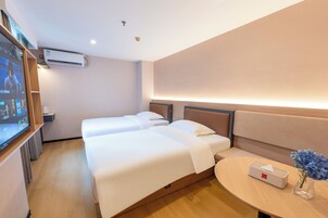 A hotel room with two beds, a bedside table with a tissue box and a vase of flowers, an air conditioning unit, and a TV screen displaying a program.