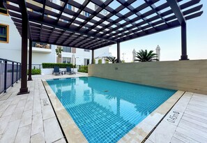 Una piscina con una pérgola sobre ella, rodeada por un solárium con baldosas y un muro bajo.