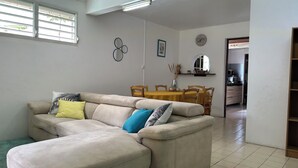 Una sala de estar con un sofá seccional color beige, una mesa de comedor con sillas, una cocina al fondo y un pasillo que conduce a otra habitación.