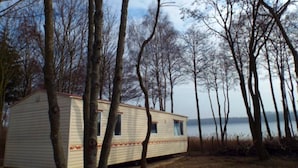 A mobile home with a small porch, surrounded by trees and a body of water in the background. - Mobile Home im Ferienpark mit Seezugang (Dabel)