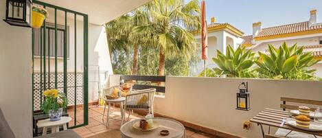 A balcony with a sofa, a dining table with a cake, a chair, a small table with a plant, and a railing.
