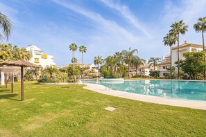 A swimming pool surrounded by grass, palm trees, and buildings with a clear blue sky. - Calanova Grand Golf (Mijas)