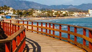 A wooden boardwalk with red railings leading to a beach with palm trees and buildings in the background.