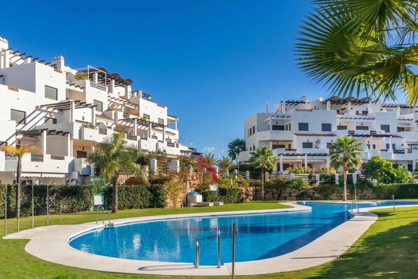 A swimming pool surrounded by white buildings with balconies, palm trees, and a clear blue sky. - Door To The Sun Estepona (Estepona)