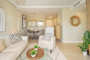 A living room with a sofa, armchair, coffee table with plants, a dining area with chairs, and a large mirror on the wall. - Door To The Sun Estepona (Estepona)
