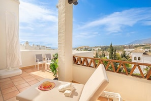 A balcony with a white chair, a small table with a hat and towel, and a view of a town with buildings and mountains. - Door To The Sun Estepona (Estepona)
