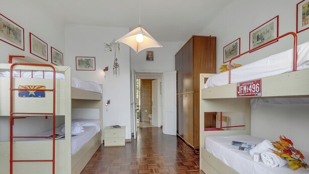 Habitación tipo dormitorio con literas, suelo de madera, lámpara colgante y una puerta que da a otra habitación.