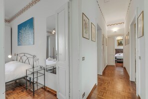 A bedroom with a bed, bedside table, and a nightstand. A hallway with wooden flooring and white walls leads to another room. - Villa Incanto Sul Lago: Lakefront, Garden & Balconies (Laveno Mombello)