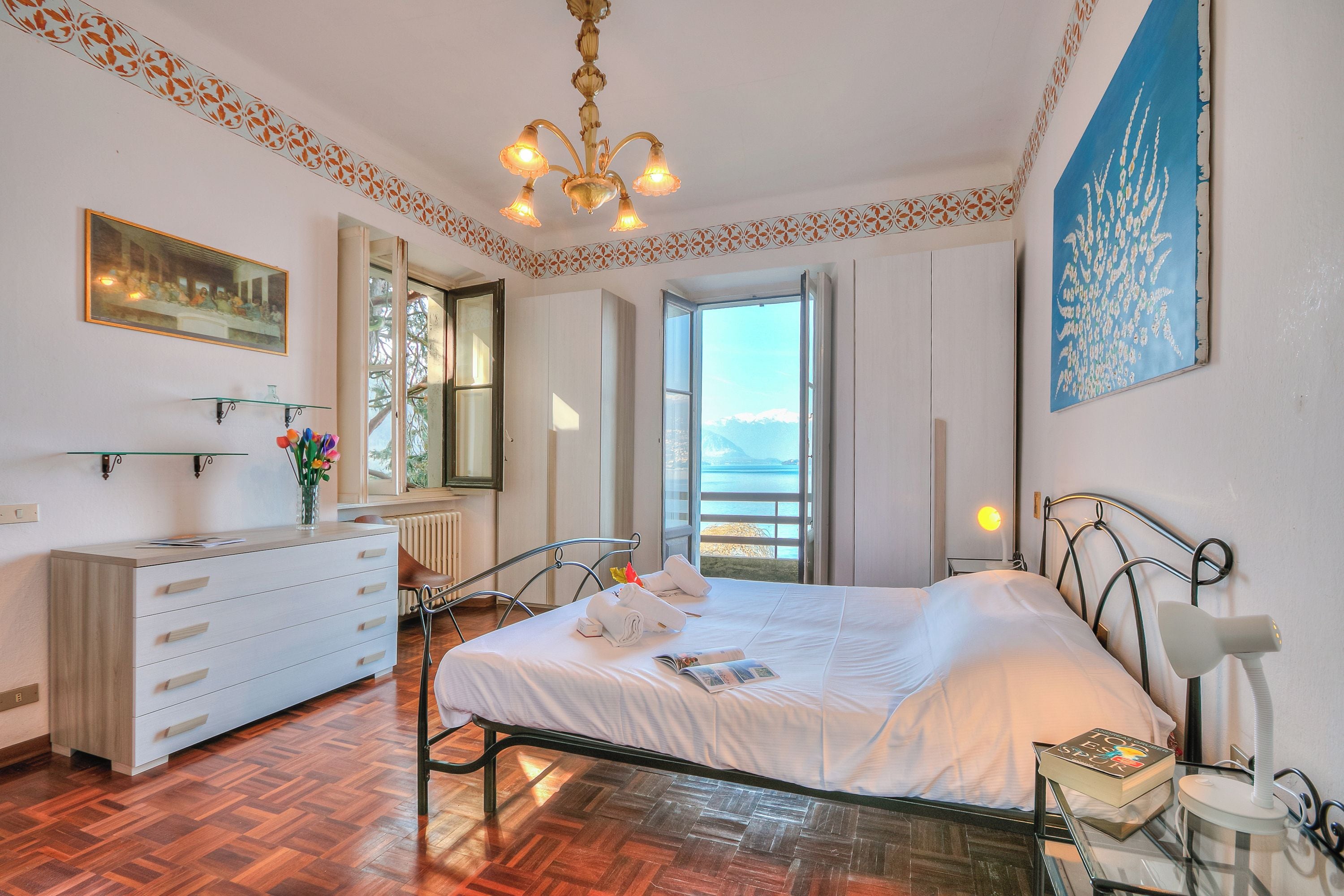 A bedroom with a bed, a dresser, a nightstand, a lamp, a painting, and a window with a view.