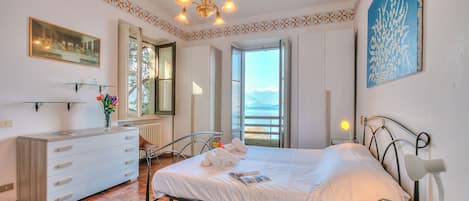 Un dormitorio con cama, cómoda, mesita de noche, lámpara, un cuadro y ventana con vistas.