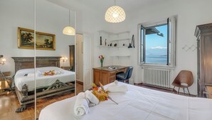 A hotel room with a large bed, a desk, a chair, a bookshelf, a mirror, and a window with a view. - Villa Incanto Sul Lago: Lakefront, Garden & Balconies (Laveno Mombello)