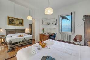 A hotel room with a large bed, a desk, a chair, a bookshelf, a mirror, and a window with a view. - Villa Incanto Sul Lago Lake Front (Laveno Mombello)
