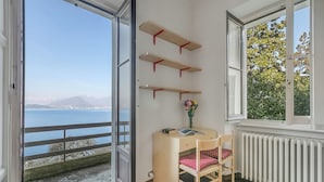 A room with a wooden desk, chair, and three shelves against the wall, a window with a view of a body of water and mountains, and a radiator below the window. - Villa Incanto Sul Lago: Lakefront, Garden & Balconies (Laveno Mombello)