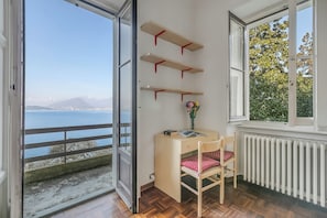 A room with a wooden desk, chair, and three shelves against the wall, a window with a view of a body of water and mountains, and a radiator below the window. - Villa Incanto Sul Lago: Lakefront, Garden & Balconies (Laveno Mombello)