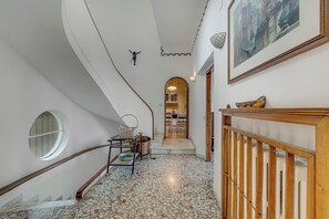 A hallway with a curved staircase, a round window, a metal rack with baskets, a copper pot, and a framed picture on the wall. - Villa Incanto Sul Lago: Lakefront, Garden & Balconies (Laveno Mombello)