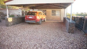 Un carport riparato con vialetto sterrato, un muro in mattoni e un edificio con finestra e porta.