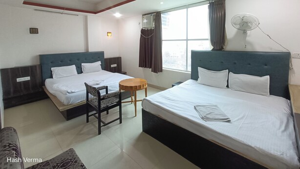 A hotel room with two beds, a small round table, a chair, a ceiling fan, and a window with curtains. - New Holiday Inn Hotel (Ajmer)