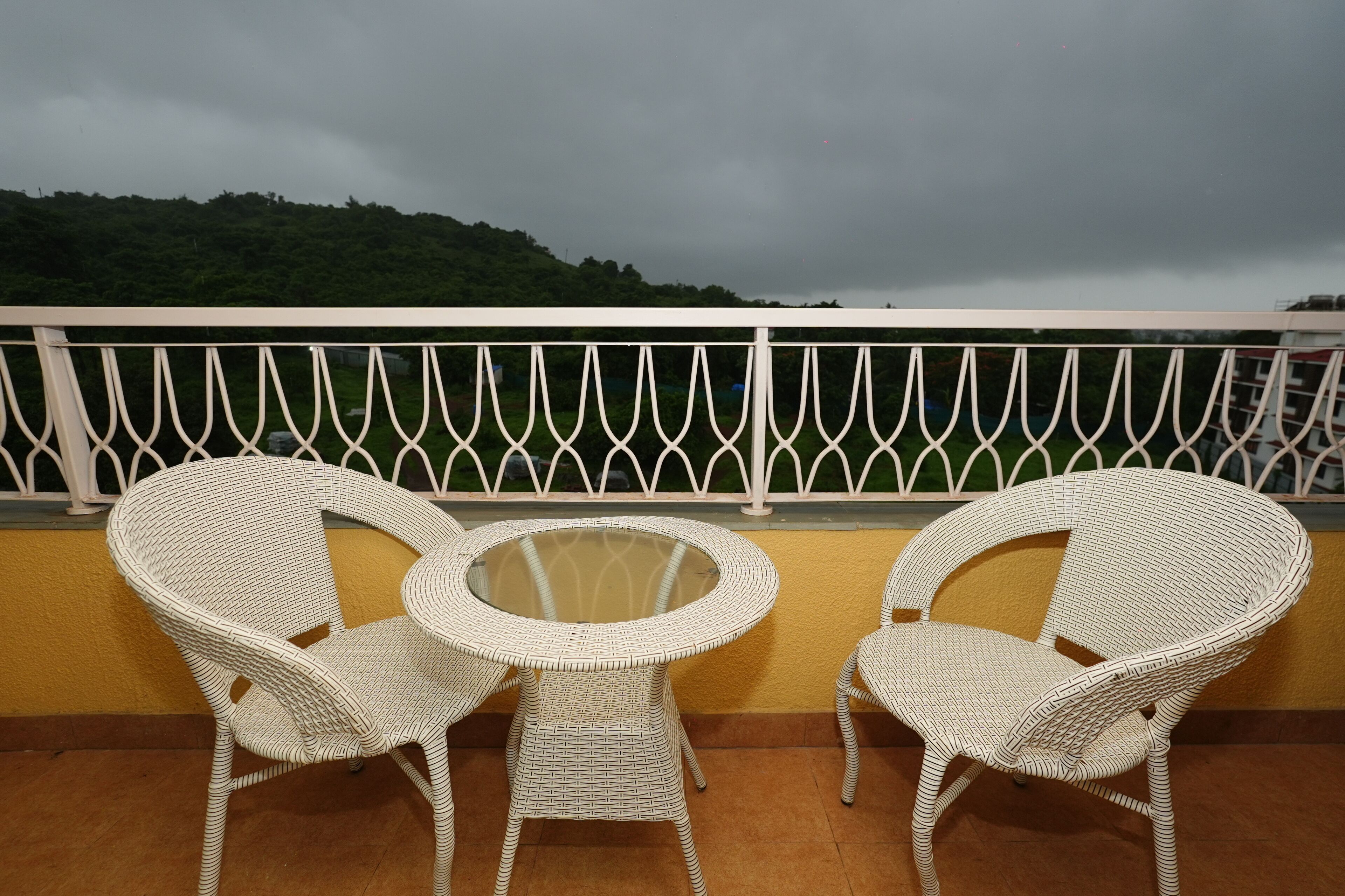 A balcony with two wicker chairs and a glass table, overlooking a lush green hillside.