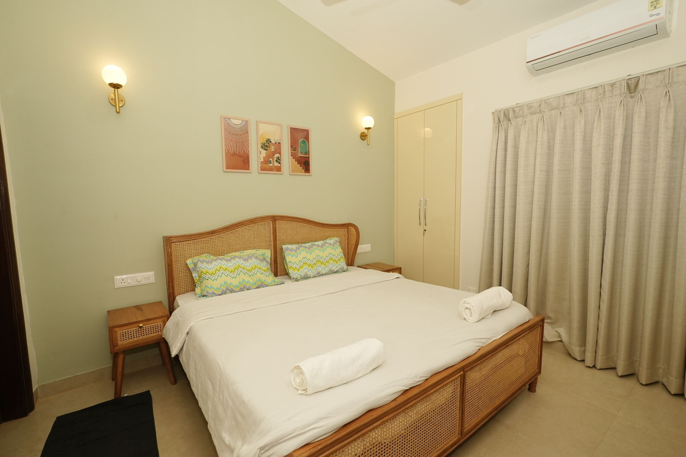 A neatly made bed with a white duvet, two pillows, and a rolled towel on the side. There are two bedside tables with lamps, a wall-mounted air conditioner, and a door with a curtain.