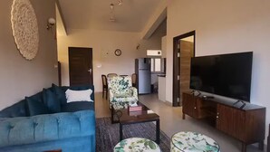 Ein Wohnzimmer mit Sofa, Poufs, Couchtisch, Fernseher und einer Küche im Hintergrund.