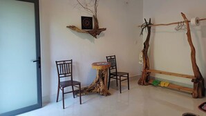Een kamer met een ronde houten tafel, twee stoelen, een wandplank met een afbeelding en een bijzondere houten constructie met een hangplant.