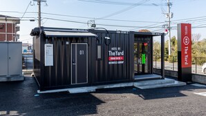 A compact hotel structure with a front desk, labeled as "The Yard" by R9 Hotels Group, featuring a simple entrance and a visible parking area.