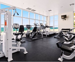 A gym with cardio machines, weight equipment, and large windows overlooking a beach. - Crystal Shores - Beachfront 2BR, Gulf Front Views, Indoor/Outdoor Pool, Hot Tub (Gulf Shores)