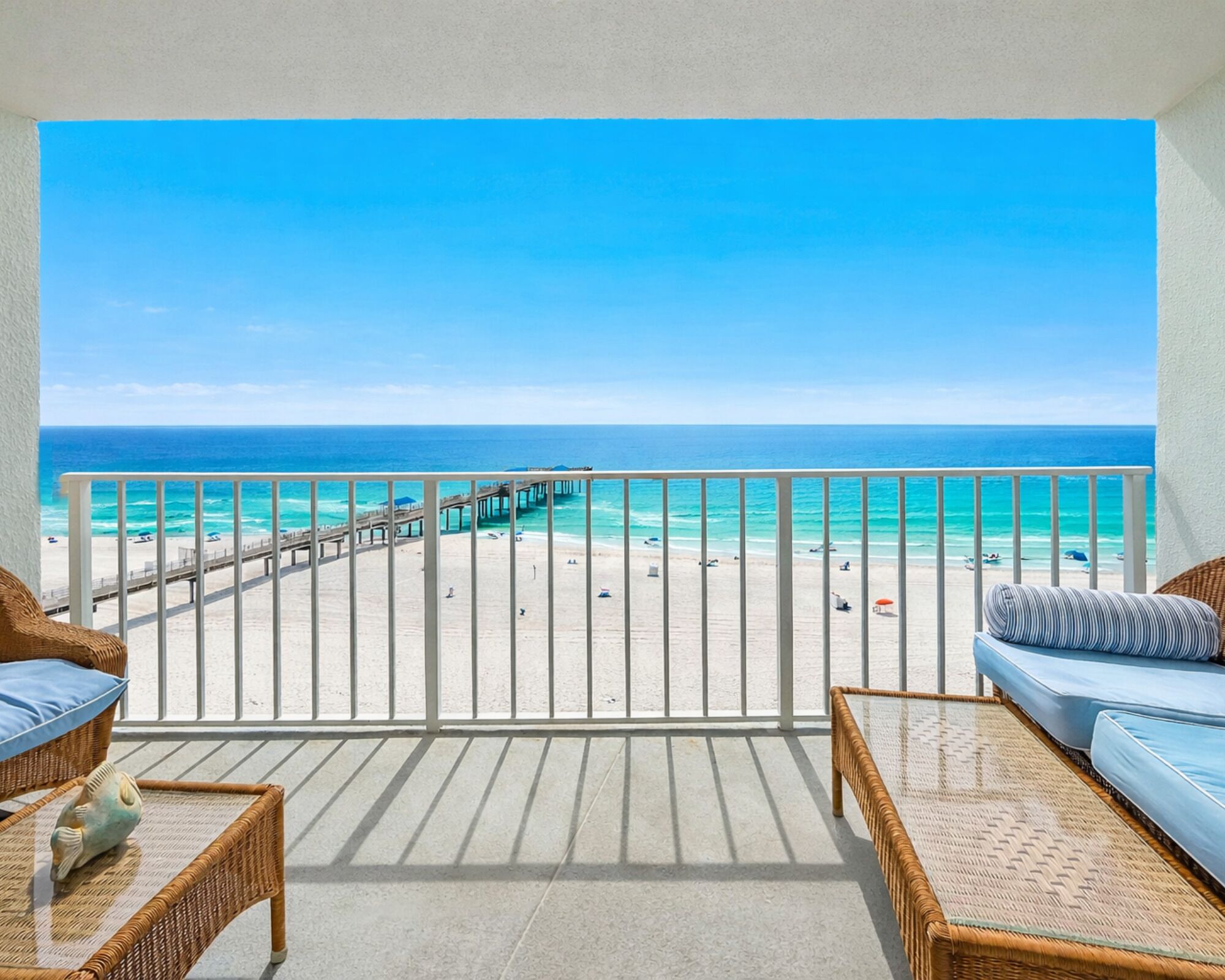 A balcony with a view of a beach and pier. There are wicker furniture and a small decorative fish.
