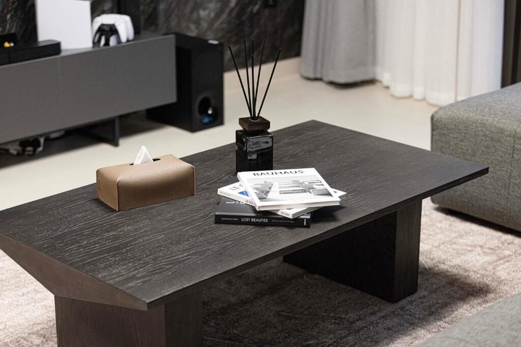 A modern living room with a dark wood coffee table, a small brown bag, a black vase with sticks, and a magazine.