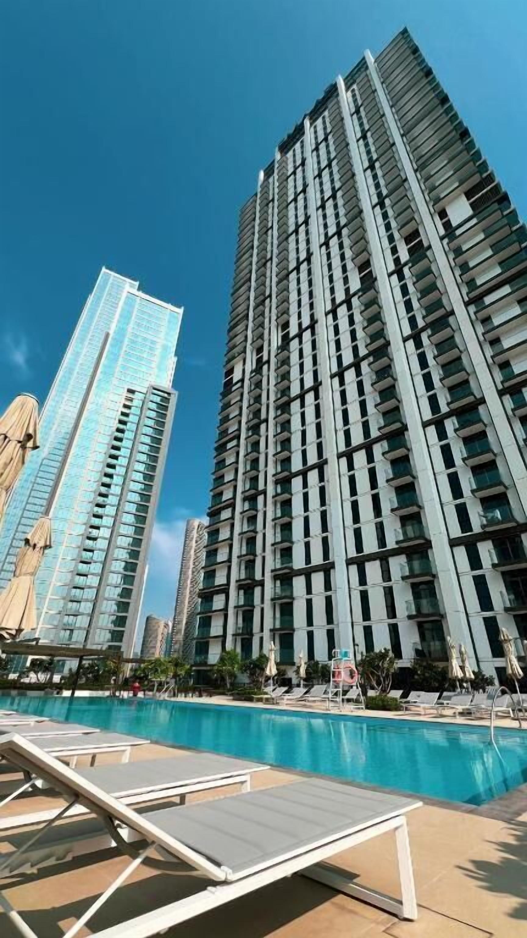 A high-rise building with a modern design, a swimming pool area with sun loungers, and a clear blue sky.