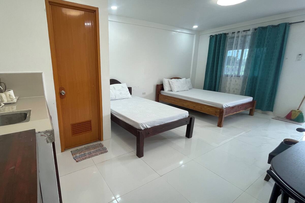 A hotel room with two single beds, a kitchenette with a kettle and mugs, a wooden door, and a bathroom with a shower curtain.