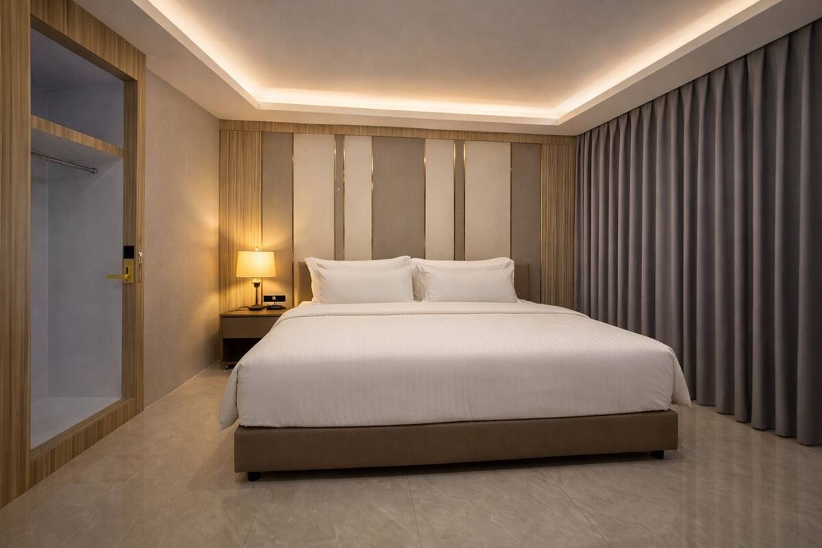 A modern hotel room with a large bed, a bedside table with a lamp, a wardrobe, and a curtain-covered window.