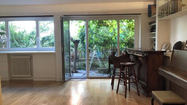 Een ruime kamer met een houten vloer, een piano, een bar met krukken en een glazen schuifdeur naar een tuin.