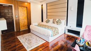 Um quarto com piso de madeira, uma cama com cabeceira branca, uma mesinha de cabeceira, uma cadeira e uma pequena mesa com objetos sobre ela.