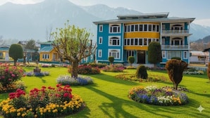 Um hotel de vários andares com fachada azul e amarela, rodeado por jardins bem cuidados e montanhas ao fundo.