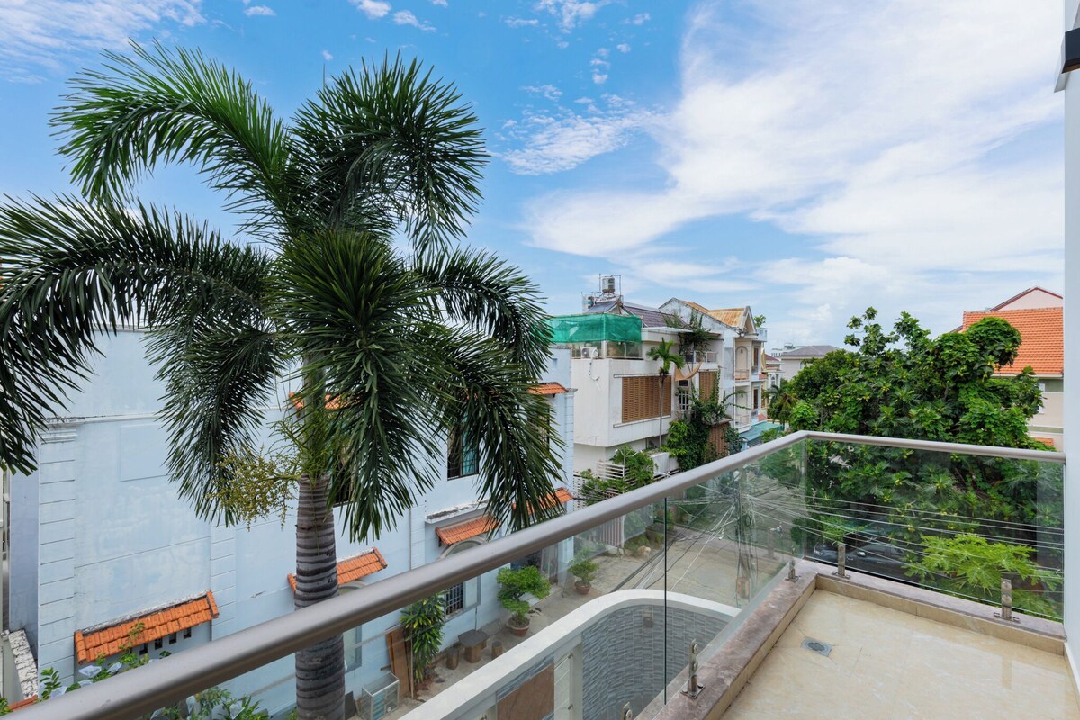 Een balkon met glazen hekwerk met uitzicht op een woongebied, met palmbomen en huizen met oranje pannendaken.