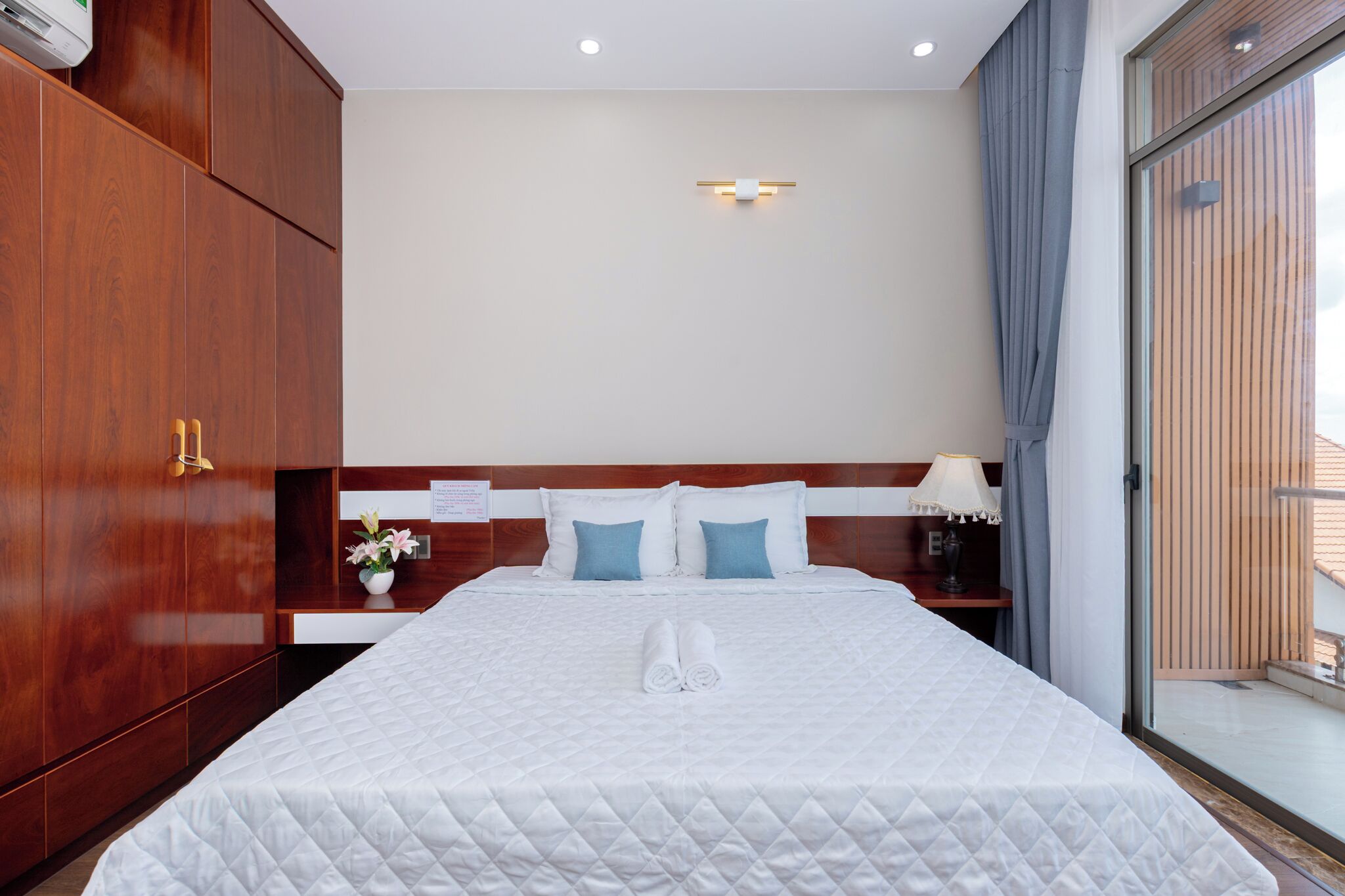 A hotel room with a large bed, wooden wardrobe, a nightstand with a lamp, and a view of the outside through a sliding door.