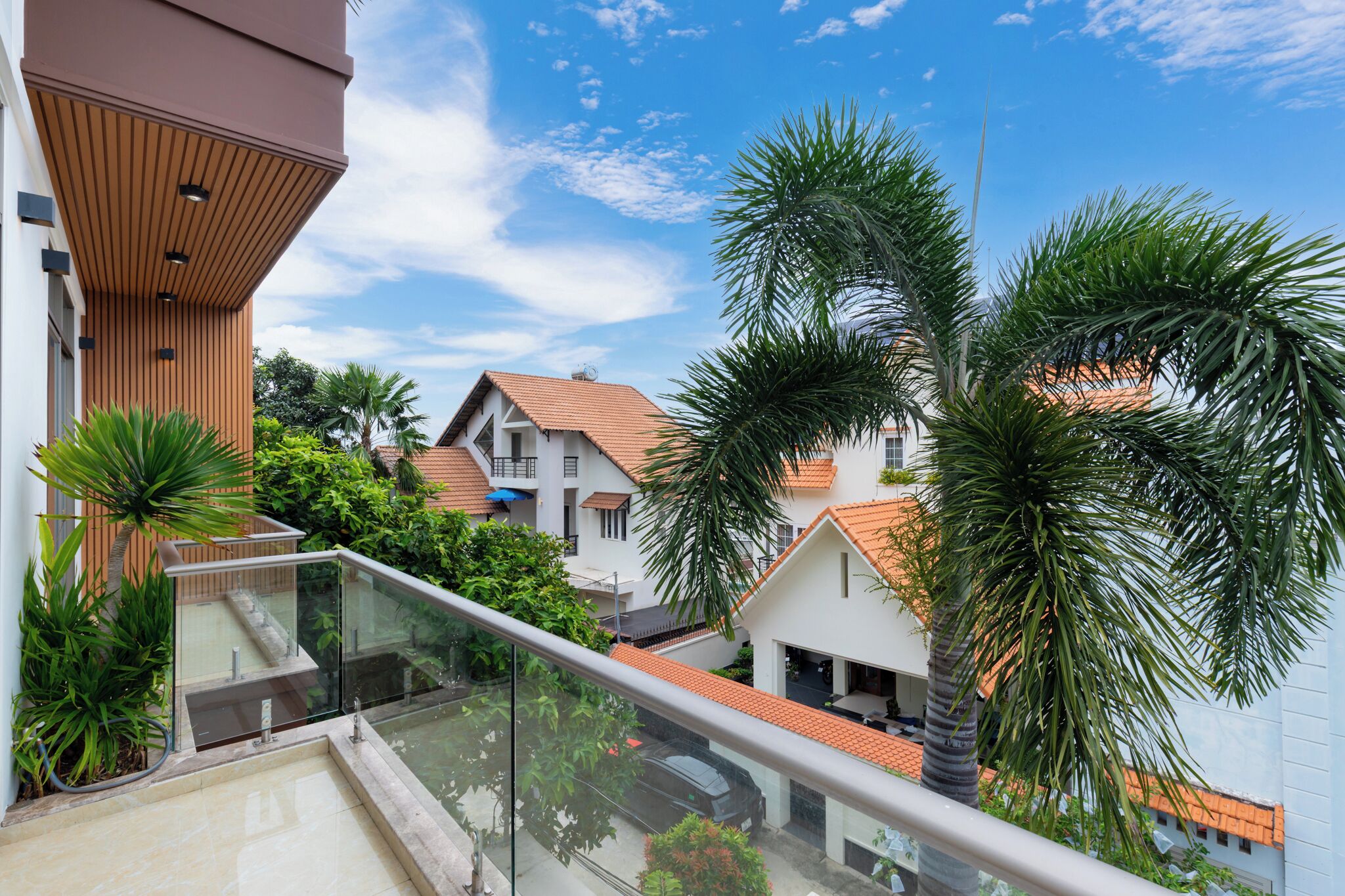 A residential area with modern houses, a balcony with glass railings, and lush greenery including palm trees.