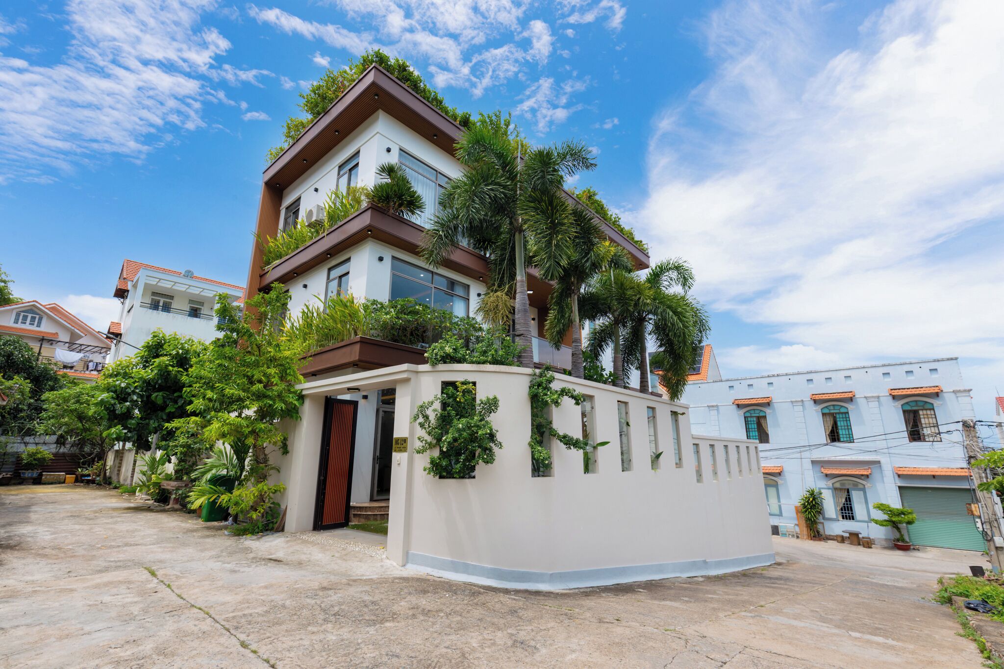 A modern two-story house with a balcony, surrounded by lush greenery and a clear blue sky.
