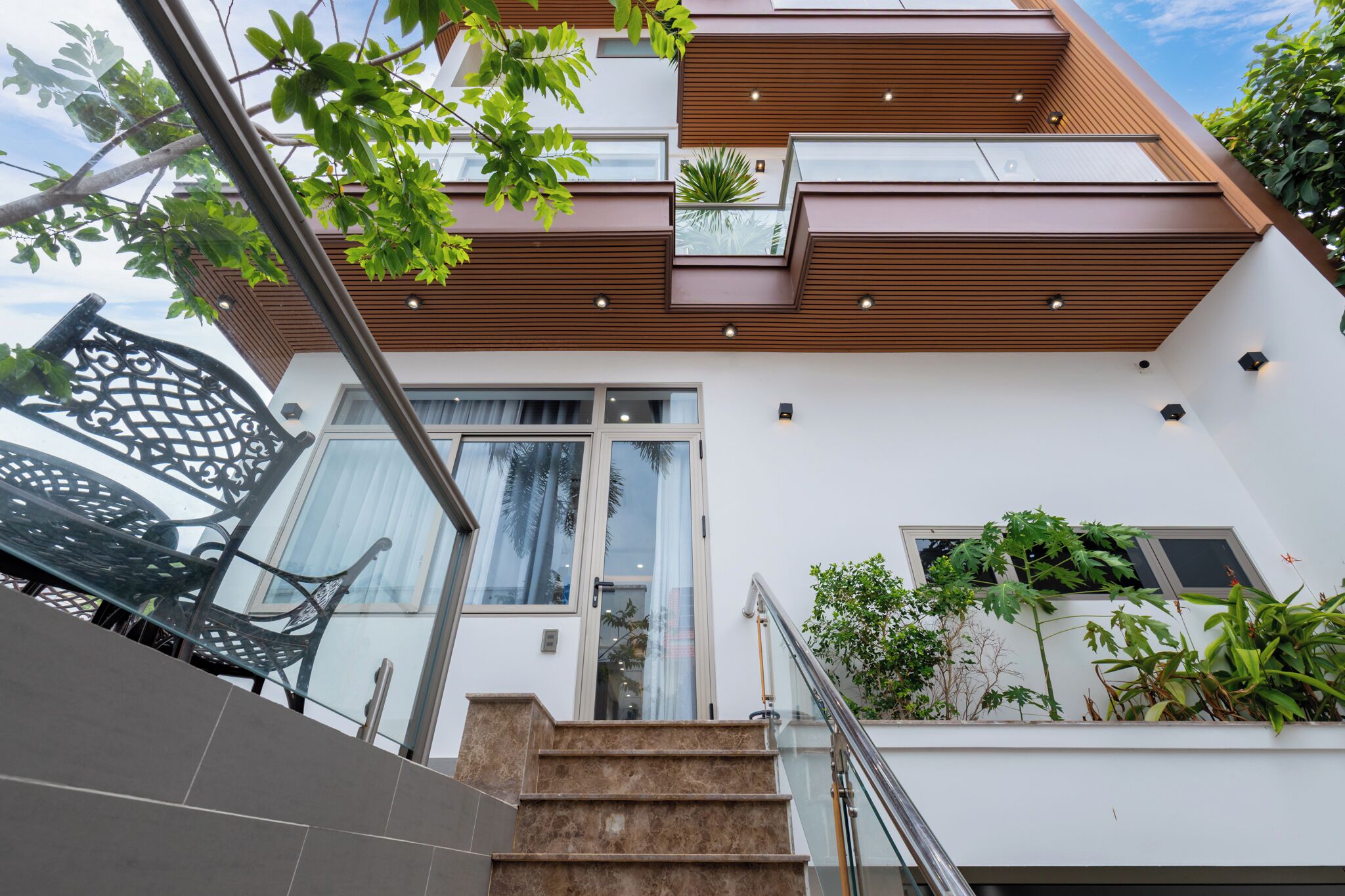 A modern building with a staircase leading to a balcony, surrounded by greenery and a glass door.