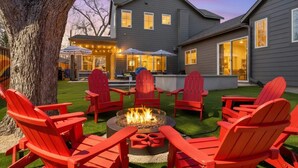 Ein Garten mit Feuerstelle, umgeben von roten Adirondack-Stühlen, einem Baum und einem Haus mit mehreren Fenstern.