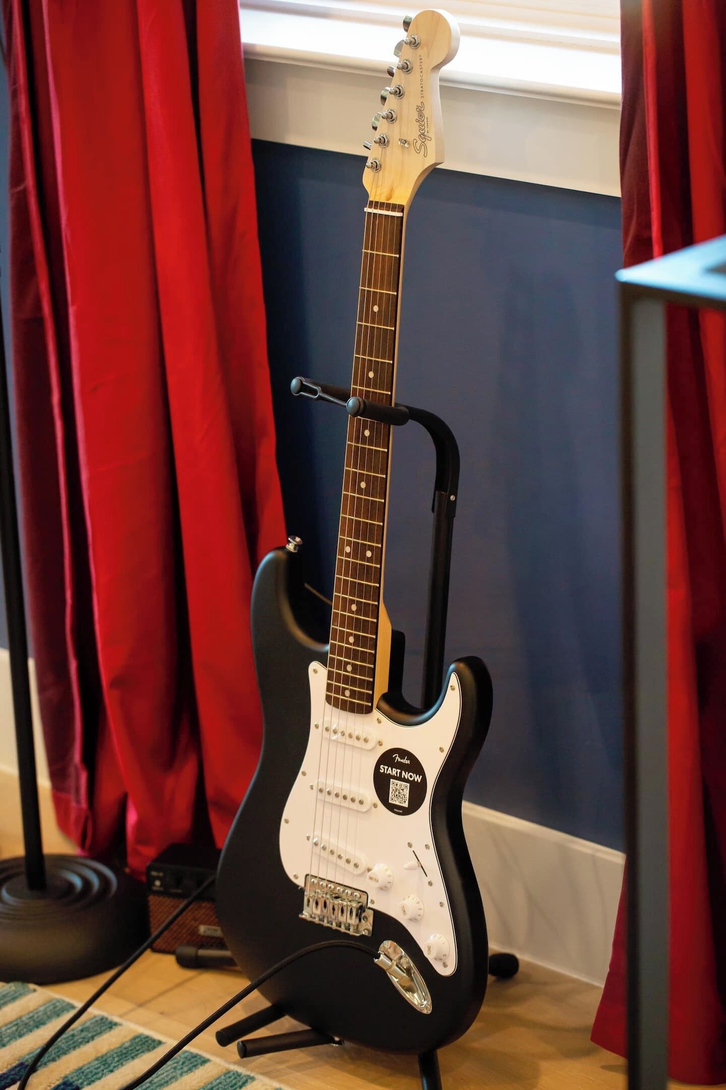 A black electric guitar with a white pickguard and a headstock with tuning pegs, placed on a stand in a room with red curtains and a blue wall.