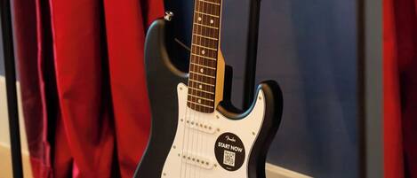 A black electric guitar with a white pickguard and a headstock with tuning pegs, placed on a stand in a room with red curtains and a blue wall.