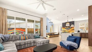 Ein modernes Wohnzimmer mit einem Ecksofa, einem Couchtisch und einem blauen Sessel. Es gibt eine Küchenzeile mit einem Esstisch und Stühlen, sowie ein großes Fenster mit Vorhängen.