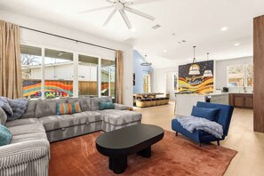 Ein modernes Wohnzimmer mit einem Ecksofa, einem Couchtisch und einem blauen Sessel. Es gibt eine Küchenzeile mit einem Esstisch und Stühlen, sowie ein großes Fenster mit Vorhängen.