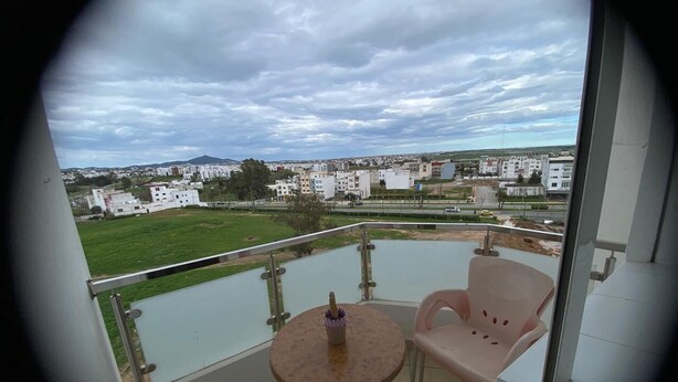 A balcony with a table and a chair, overlooking a residential area with buildings and a river. - Luxury Apartment with Mountain View and Parking in Tetouan (Tétouan)
