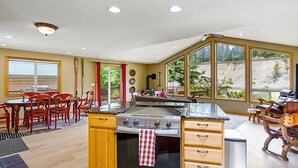 A kitchen with a stove, wooden cabinets, a dining area with chairs, and large windows. - Prospector's Bend - Hot Tub, Riverfront, Fishing (Gold Bar)