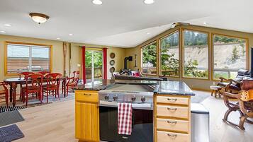 A kitchen with a stove, wooden cabinets, a dining area with chairs, and large windows.